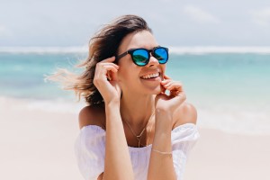 Wonderful woman in white attire and sparkle glasses posing with happy face expression in hot summer day. Pleasant caucasian girl standing near ocean on sky background..